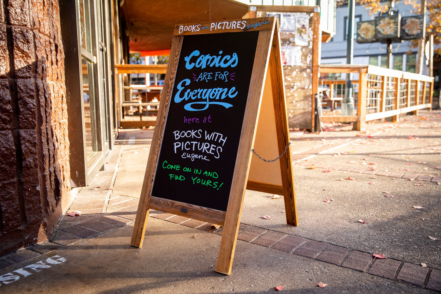 wooden sandwich board with chalk writing reading Comics Are For Everyone at Books With Pictures Eugene on a sunny sidewalk next to red bricks