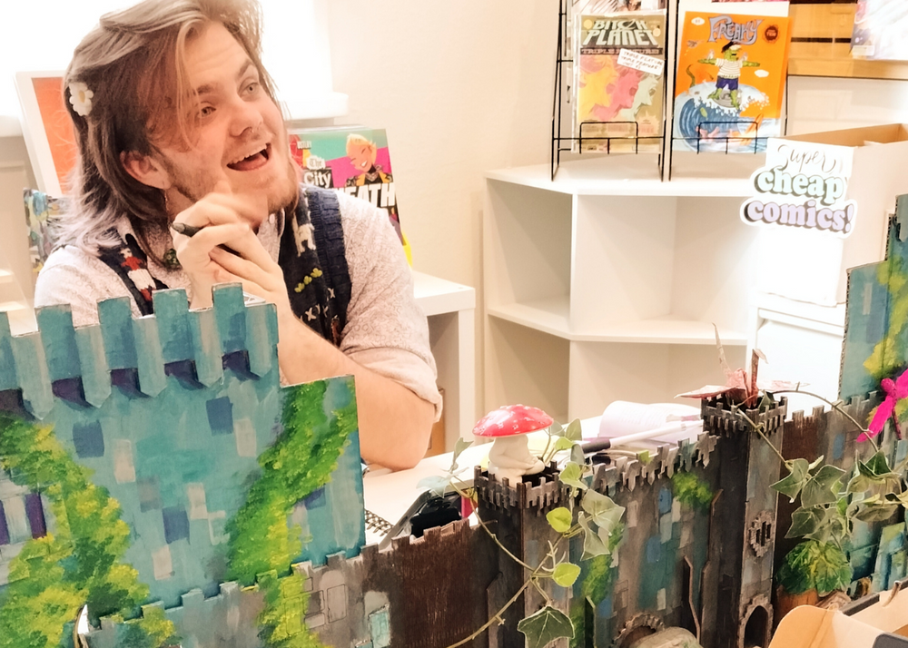A person sitting behind a large, whimsical castle diorama made of painted wood, with green ivy, mushrooms, and fantasy scenery, in a brightly lit room with shelves and books in the background.
